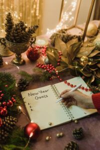 Hand writing New Year goals in a festive notebook surrounded by Christmas decorations and fairy lights indoors.